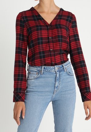 Punainen ja laivastonsininen ruudullinen paita, jossa on V-aukko ja pitkät hihat, yhdistetty vaaleansinisiin korkeavyötäröisiin farkkuihin. Sileä kangaspinta.