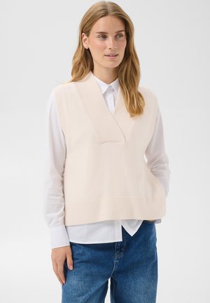 Femme aux longs cheveux blonds portant un gilet en tricot sans manches couleur crème par-dessus une chemise blanche à boutons et un jean bleu, debout devant un fond clair.