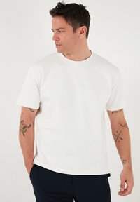 Camiseta blanca de cuello redondo, de mangas cortas, con textura suave. Llevada con pantalones oscuros, con tatuajes visibles en los brazos.