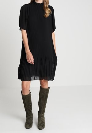 Woman wearing a knee-length black pleated dress with short sleeves and olive green suede knee-high boots.