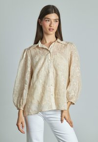 Blusa color beige chiaro con ricami floreali, apertura con bottoni sul davanti e maniche a sbuffo. Realizzata in tessuto trasparente e testurizzato. Abbinata a pantaloni bianchi.