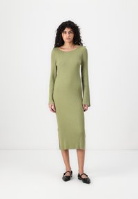 Ribbad, långärmad klänning i olivgrönt med sidofickor, figursydd silhuett och bred neck-line, kombinerad med svarta ballerinat.