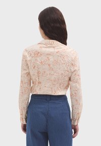 Camicia beige con illustrazioni arancioni, con maniche lunghe e colletto classico. Indossata con pantaloni navy a righe.