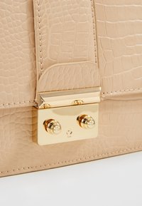 Beige läderhandväska med krokodiltextur, med ett lås och hårdvara i guldton, rundade detaljer och stickade kanter.