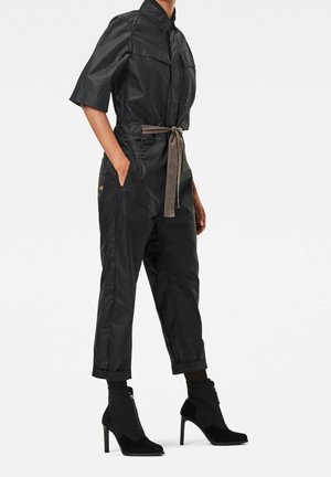 Musta jumpsuit, jossa on lyhyet hihat, vyötäröllä vyö, kaksi rintataskua ja käännetyt hihansuut, yhdistetty mustiin mokkanahkaisiin nilkkureihin, joissa on vetoketju.