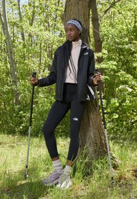 Crna najlon jakna, svijetlo ružičasta majica, crne tajice, bijeli logotip. Sive planinarske cipele s bijelim prugastim čarapama, planinarski štap, pozadina sa zelenim lišćem.