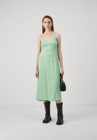 Vestido midi verde con un patrón de flores blancas, tirantes finos y un corsé ajustado, combinado con botas negras hasta la rodilla y un pequeño bolso negro.