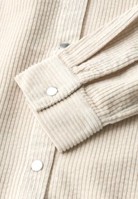 Close-up van een crèmekleurige corduroy jas met mouwen en voorzijde, met zilveren drukknoopsluitingen, waarbij de textuur en sluitdetails duidelijk in beeld worden gebracht.
