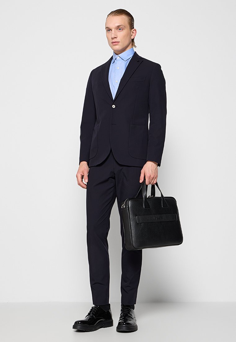 Schwarze Lederaktentasche mit doppelten Griffen, kombiniert mit einem marineblauen Anzug und einem hellblauen Hemd, das einen Einzelknopf und schmal geschnittene Hosen hat.