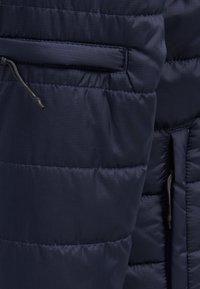 Tejido de chaqueta puffer azul marino con secciones acolchadas y una textura suave. Presenta un bolsillo con cremallera y ajuste con cordón en el dobladillo.