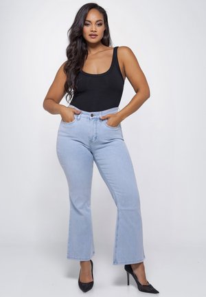 Vrouw met lang donker haar, gekleed in een zwart mouwloos topje, lichtblauwe hoge taille jeans en zwarte hoge hakken, poserend met handen in de zakken.