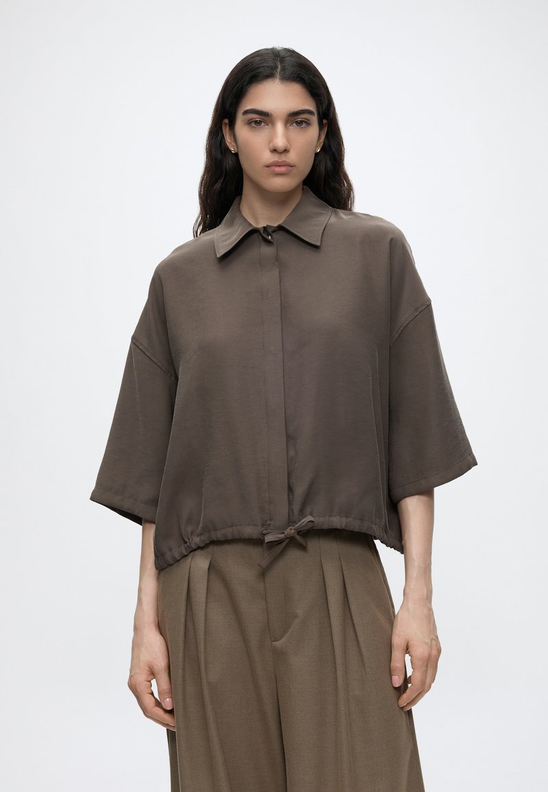 Femme aux longs cheveux foncés portant une chemise marron ample à manches courtes avec un cordon à la taille et un pantalon marron large assorti.