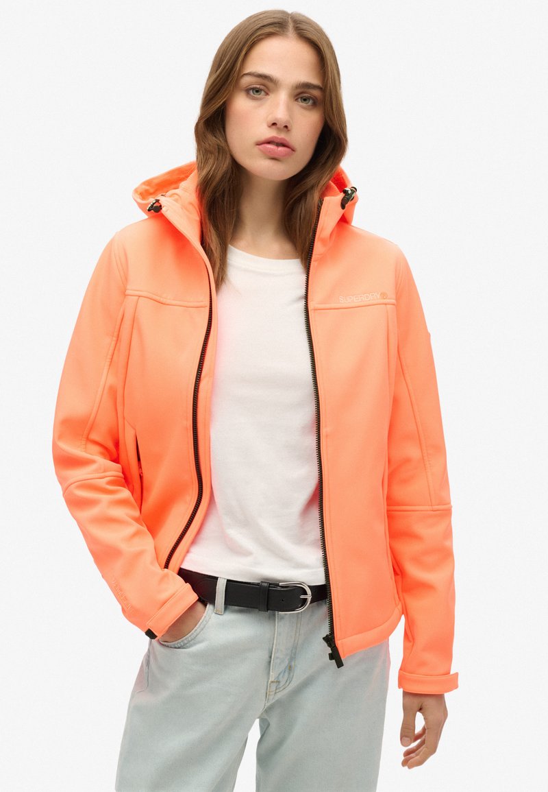 Veste à fermeture éclair orange néon avec capuche, présentant un tissu lisse et brillant, des accents de fermeture éclair noirs, et un design ajusté associé à une chemise blanche.
