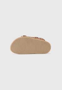 Sandal med perforert design i lysebrun, med en flat, stripete gummisåle. Oversiden viser utskårne mønstre for ventilasjon.