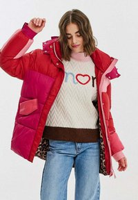 Žena v červené a růžové péřové bundě přes bílý svetr s barevným nápisem, doplněná o modré džíny, na jednobarevném pozadí.