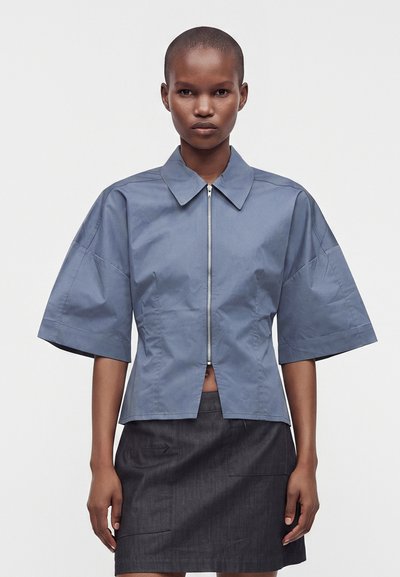 Personne noire aux cheveux courts porte une chemise bleu clair à manches courtes avec une fermeture éclair et une jupe gris foncé, debout devant un fond blanc uni.