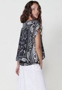 Haut en imprimé paisley noir et blanc avec des manches courtes et un ourlet froncé. Texture lisse, design fluide, porté avec un pantalon blanc.