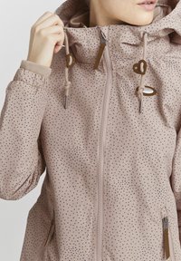 Giacca rosa chiaro con piccoli pois scuri, chiusura con zip, polsini elasticizzati, cordini e dettagli in pelle decorativi sulla cappuccio e sulla zip.