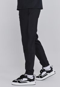 Schwarze Jogginghosen mit elastischen Bündchen, leichtem Material und tonal geprägtem Branding auf dem Oberschenkel, kombiniert mit schwarz-weißen Sneakers.