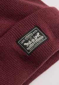 Bordeaux strikket hue med tekstureret mønster, udstyret med et sort rektangulært mærke med hvidt logo og tekstdetaljer. Blødt materiale.