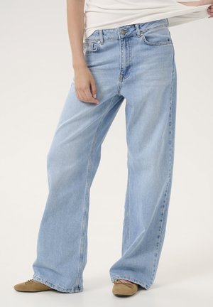 Osoba ubrana w jasnoniebieskie spodnie o szerokich nogawkach, beżowe płaskie buty i biały top, stojąca z jedną ręką lekko odsuwającą koszulę.