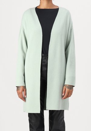 Personne portant un long cardigan vert clair ouvert sur un haut noir et un pantalon en cuir noir, montrant des tatouages sur la main.