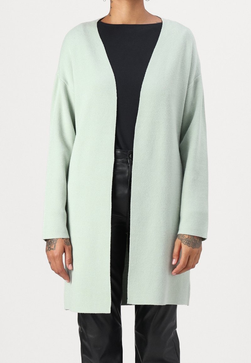 Personne portant un long cardigan vert clair ouvert sur un haut noir et un pantalon en cuir noir, montrant des tatouages sur la main.