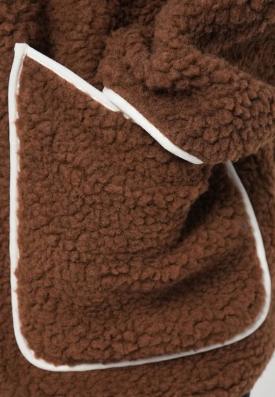 Tela de forro marrón con una superficie texturizada, que cuenta con un borde blanco contrastante a lo largo de los bordes. Un bolsillo visible está integrado en el diseño.