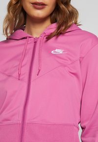 Rosa zip-up hoodie med en känguruficka, justerbar dragkedja och vit Nike-logotyp på vänster bröst. Slät tygtextur.