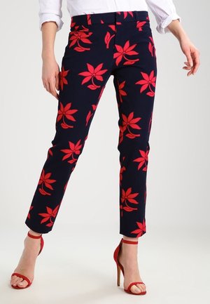 Marineblaue, schmal geschnittene Hose mit kräftigem rotem Blumenmuster, getragen zu weißem Hemd und roten Sandalen mit hohem Absatz.