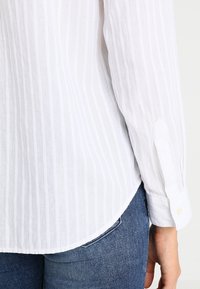 Hvid langærmet skjorte med lodrette striber, stikket ned i blå denim jeans, som viser knappen på ærmet og en del af baglommen.