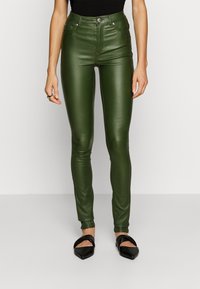 Kvinna som bär högmidjade slim-fit gröna läderbyxor och svarta spetsiga slip-on-skor, stående mot en enkel ljus bakgrund.