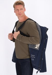 Mornarska in siva jakna s flis podlogo, zapiranje na pritisne gumbe in zadrgo, ter stranskima žepoma. Model nosi dolgo sleeved olivno majico pod njo.