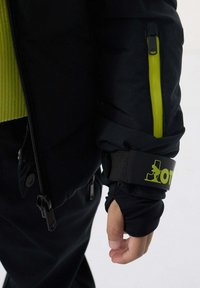 Chaqueta acolchada negra con una cremallera verde vibrante, que presenta un grueso puño negro y una correa ajustable con un logotipo, tela texturizada.