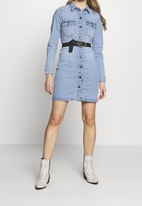 Vestido camisero de denim con mangas largas, cierre de botones en la parte delantera, dos bolsillos en el pecho y un cinturón negro en la cintura. Combinado con botas de tobillo blancas de piel de serpiente.