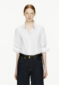 White long-sleeve button-up shirt with rolled sleeves, paired with dark denim high-waisted jeans and a black belt featuring a gold buckle.