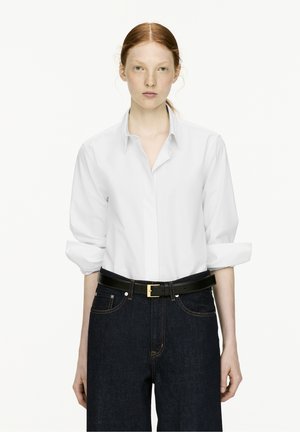 Weiße Longsleeve-Hemd mit umgeschlagenen Ärmeln, kombiniert mit dunkelblauen, hochsitztenden Jeans und einem schwarzen Gürtel mit goldener Schnalle.