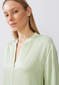 Blusa verde chiaro con colletto, realizzata in tessuto liscio, dotata di una patta e maniche leggermente arricciate, che presenta una vestibilità comoda e linee pulite.
