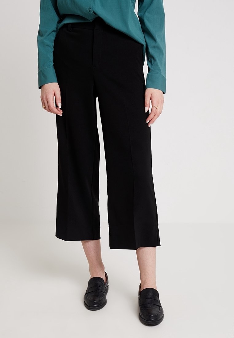 Femme portant un pantalon noir ample coupé, des mocassins noirs et une chemise boutonnée à manches longues bleu sarcelle, debout sur un sol blanc.