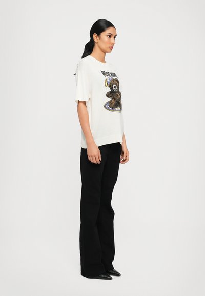 T-shirt crema a maniche corte con una grafica di orso testurizzata, abbinata a pantaloni neri a palazzo e scarpe nere a punta.