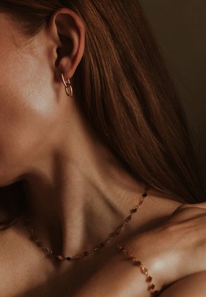 Gold-tone metal earrings featuring a linked design; delicate gold necklace with small, faceted beads; smooth skin and flowing hair are visible.