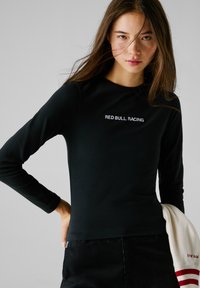 Zwart langharig shirt van gladde stof, met "RED BULL RACING" geborduurd in het wit. Model houdt een witte sweater vast.