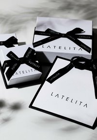 Tres cajas de regalo blancas: una grande y dos pequeñas. Cada una presenta una cinta negra con el texto "LATELITA". Las cajas tienen una textura suave y líneas limpias.