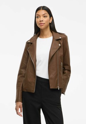 Femme portant une veste de motard en cuir brun sur un haut blanc et un pantalon noir, se tenant avec une main dans la poche devant un fond uni.
