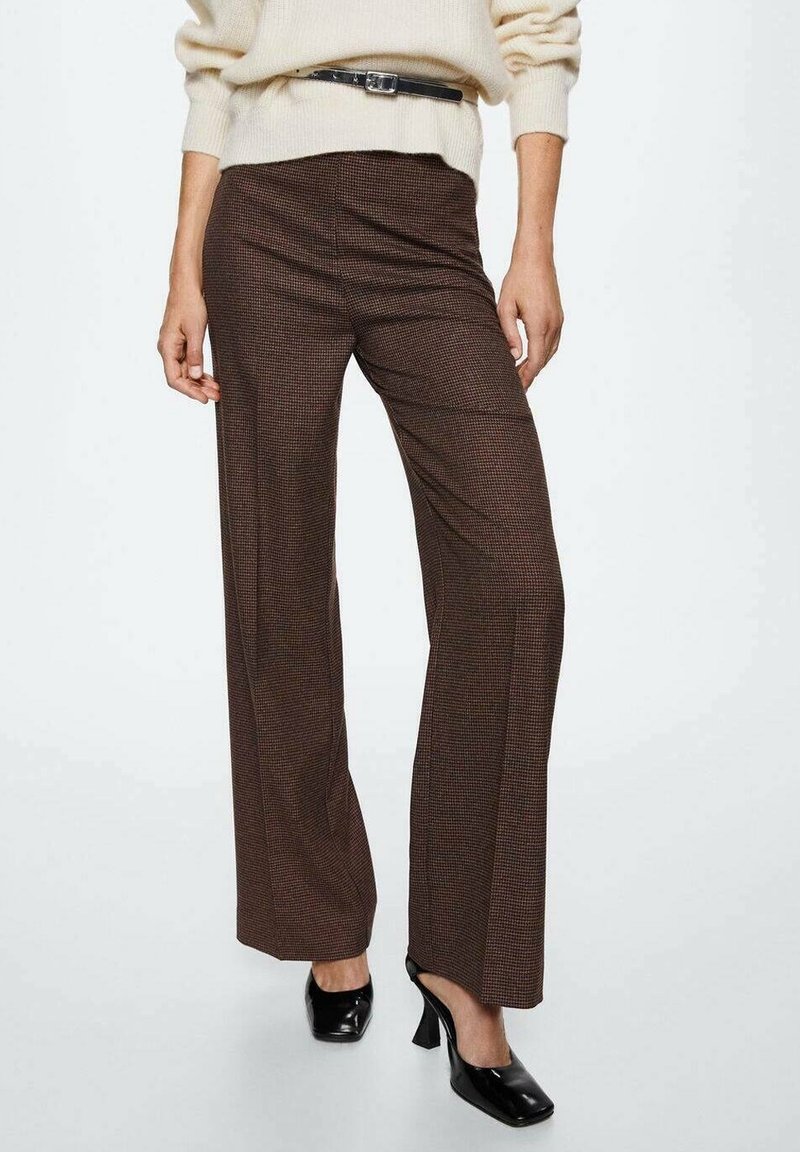 Femme portant un pantalon large à carreaux marron, un pull crème avec une ceinture noire, et des talons carrés noirs sur un fond blanc.