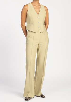 Mujer con chaleco beige sin mangas abotonado, pantalones anchos de talle alto a juego y zapatos puntiagudos con estampado de cebra.