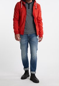 Sweat à capuche rouge zippé avec une doublure de capuche bleu marine, associé à un polo rayé et un jean en denim bleu. Des bottes noires à lacets complètent le look.