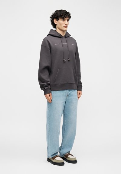 Jaunas vyras stovi, dėvi tamsiai pilką sportinį megztinį su kapišonu, šviesiai mėlynas džinsus ir rudai smėlio spalvos laisvalaikio batus priešais paprastą šviesų foną.