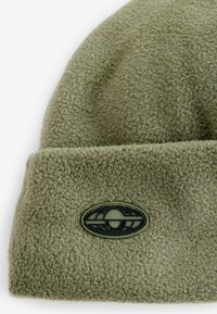 Grön fleecehatt med uppvikt mudd, som har en cirkulär svart logotyp patch sydd på framsidan. Mjuk textur med plyschfinish.