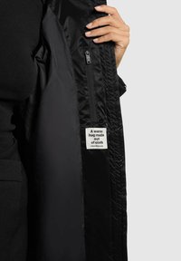 Zwarte geïsoleerde jas met een gladde buitenkant, voorzien van een ritszak en een geweven label met de tekst: "Een warme knuffel van stof."